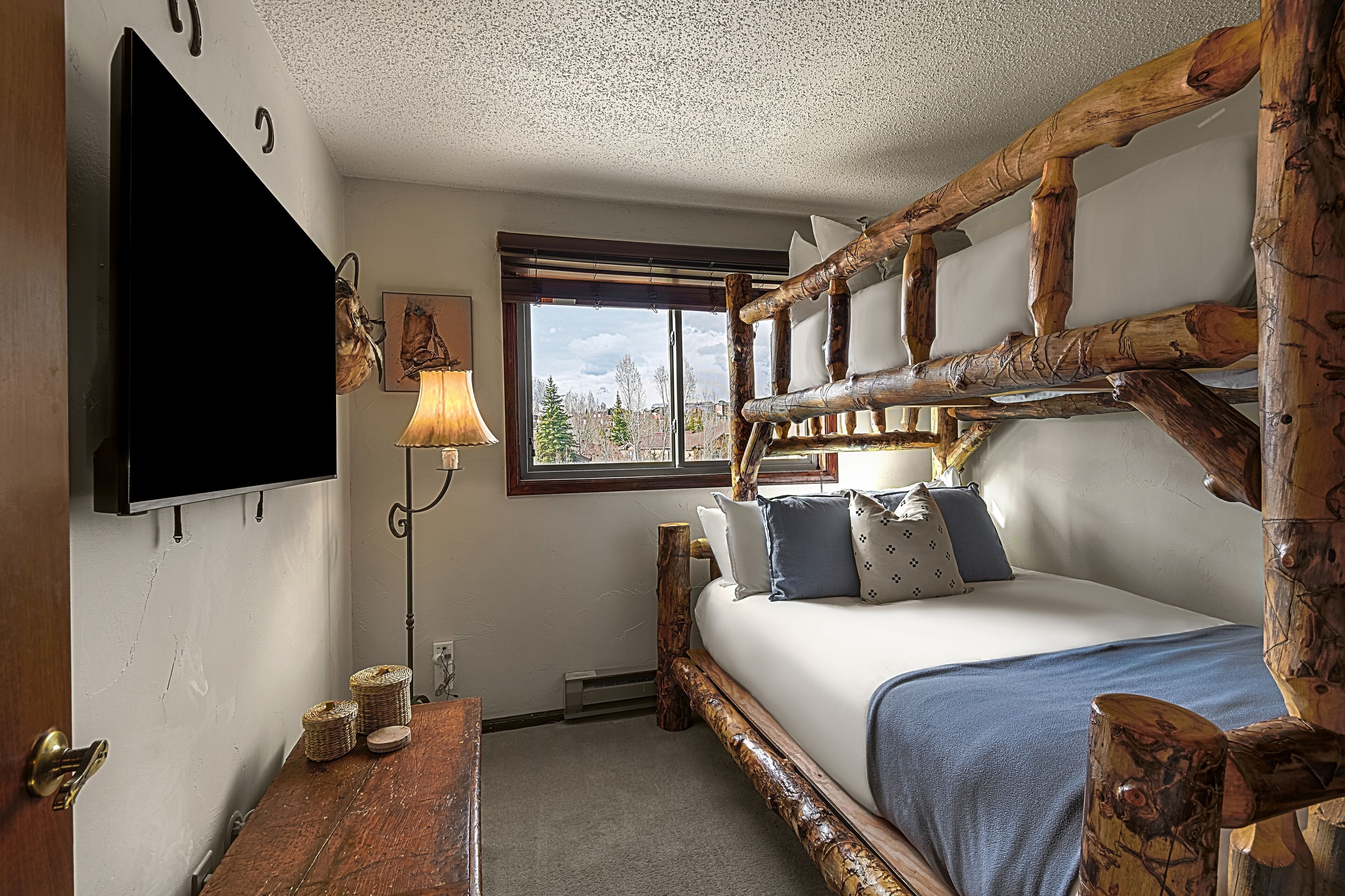 A rustic room with a wooden bed frame, a flat-screen TV mounted on the wall, a lamp, and a window with a view of trees.