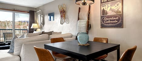 Un comedor con una mesa, sillas y una araña. Hay un sofá y una ventana con vistas. Un cartel de bienvenida está en la pared.