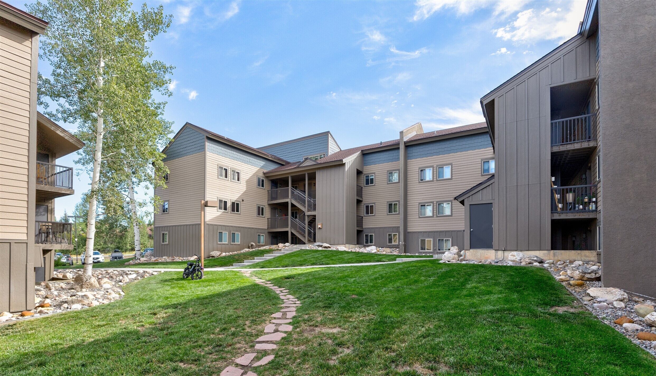 A multi-story residential building with balconies, a stone pathway, and a grassy area.