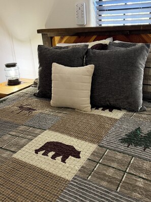 Ein Bett mit gesteppter Decke, ein Nachttisch mit Lampe und eine Steckleiste an der Wand.