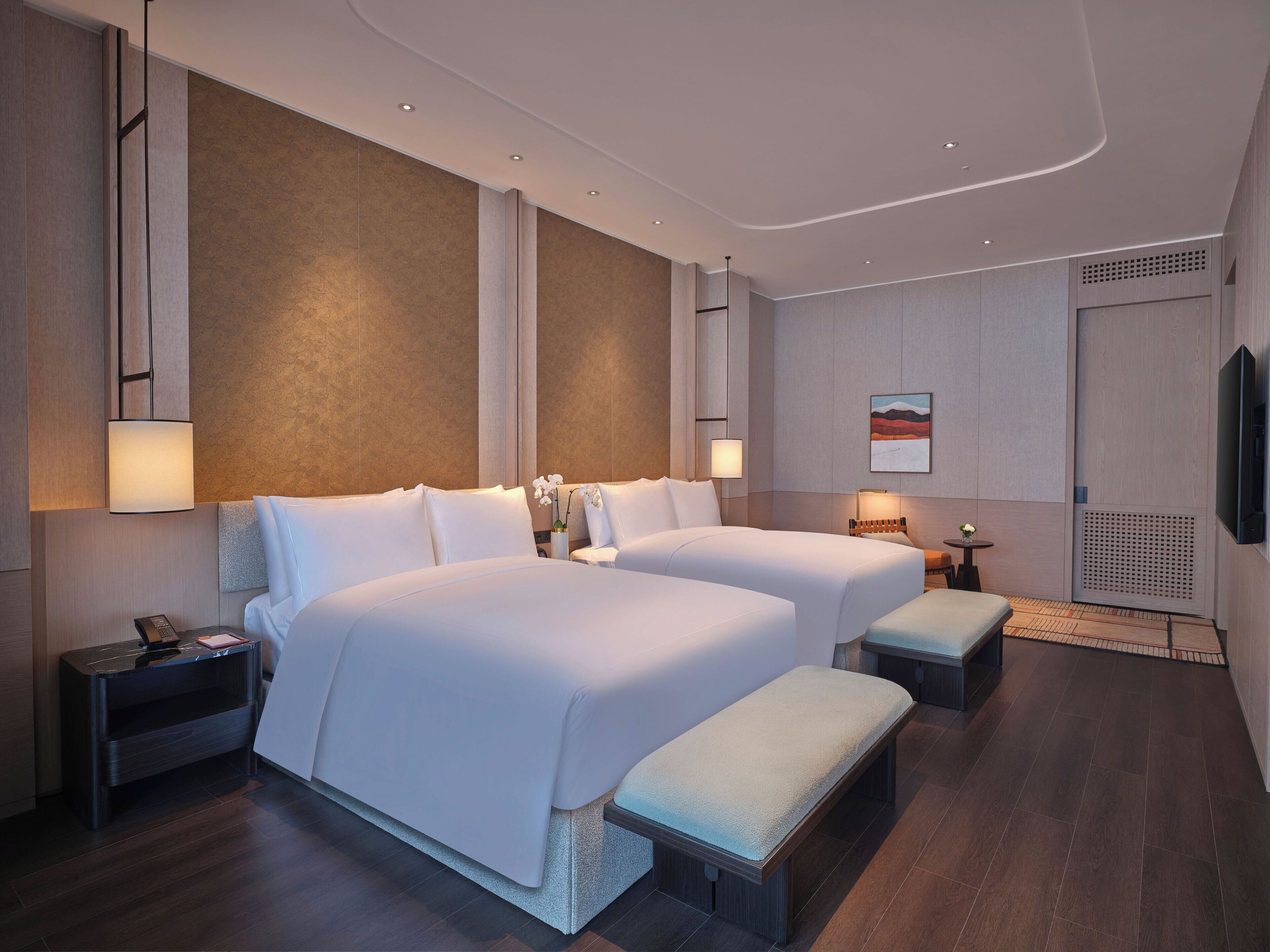 Ein modernes Hotelzimmer mit zwei Betten, Nachttischen, einem kleinen Tisch mit Lampe und einem an der Wand montierten Fernseher.