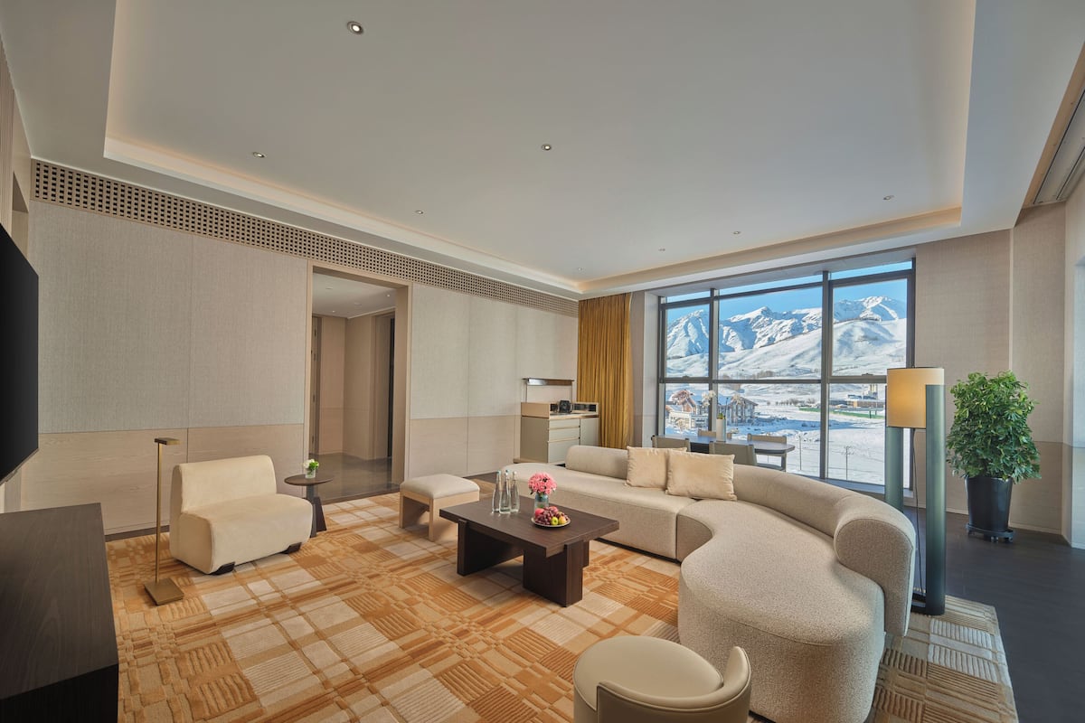 Una habitación de hotel moderna con un ventanal que ofrece vistas a la montaña, un rincón de descanso acogedor con sofá y sillones, un televisor de pantalla plana y una pequeña cocina.