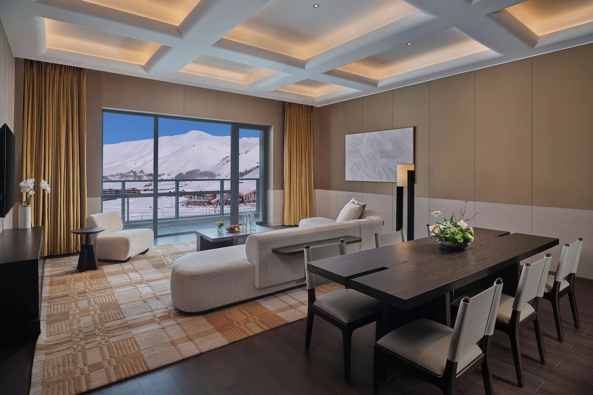 Una habitación de hotel moderna con un ventanal que ofrece vistas a la montaña, un rincón de descanso acogedor y una zona de comedor con una mesa y sillas de madera oscura.