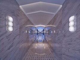 A modern, well-lit hallway with marble walls and a reflective floor, leading to a bright, open space.