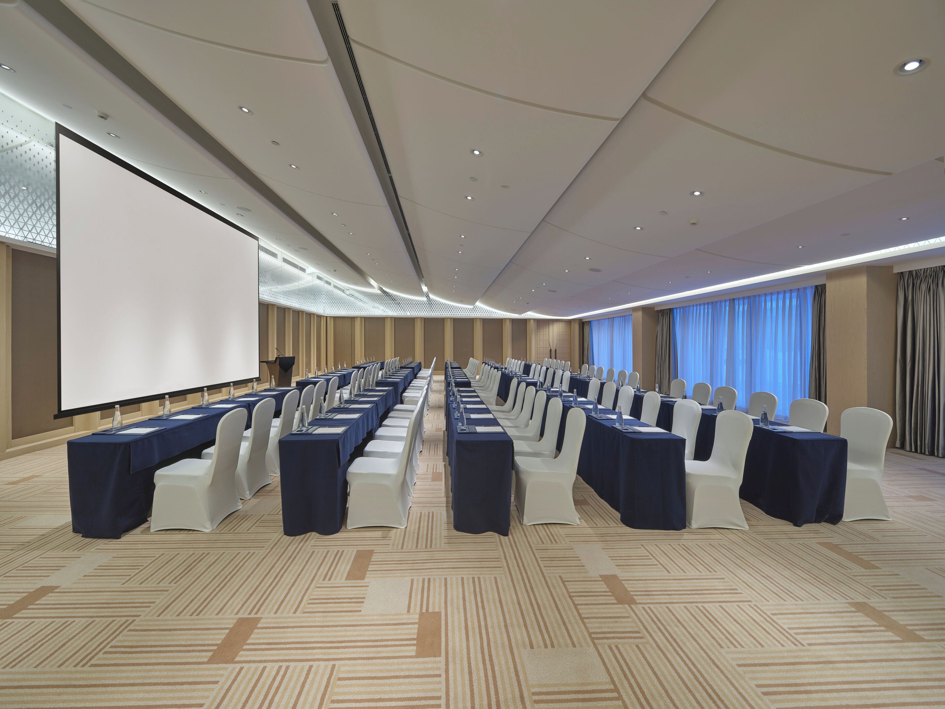 A conference room with rows of tables and chairs, a large screen, and a carpeted floor.