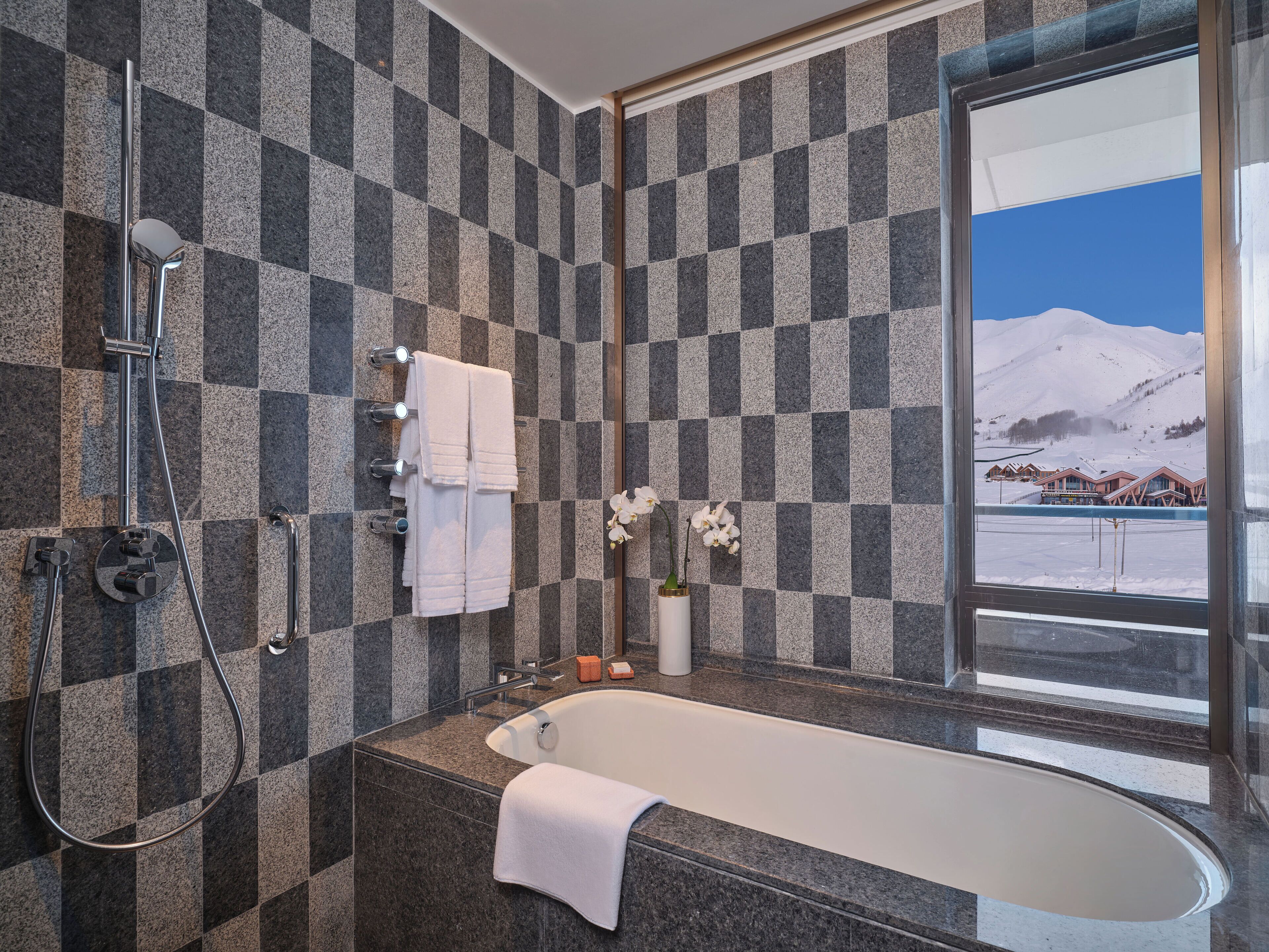 Ein Badezimmer mit Wand aus Schachbrettfliesen, einer Badewanne, einer Dusche und einem Fenster mit Blick auf einen verschneiten Berg.