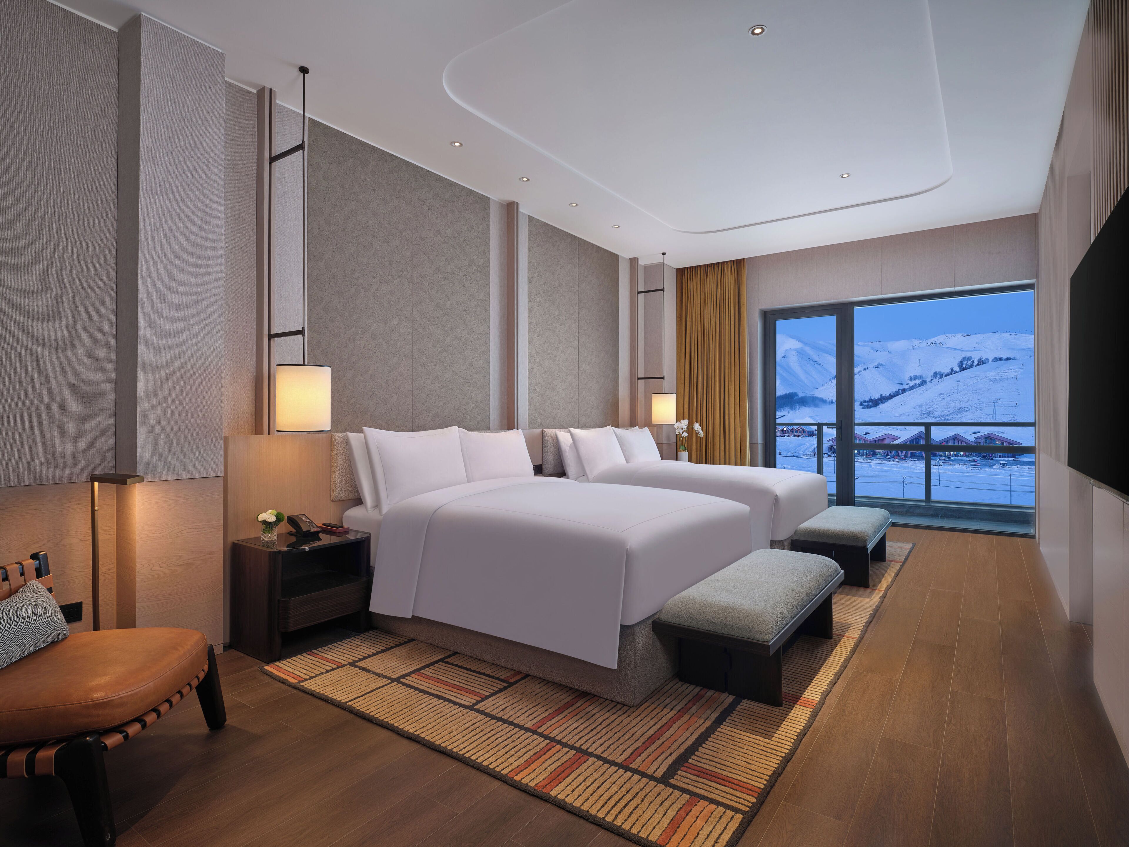 A modern hotel room with two beds, a large window with a snowy view, a wooden floor, and a small seating area.