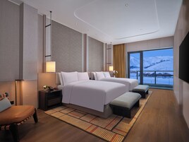 A modern hotel room with two beds, a large window with a snowy view, a wooden floor, and a small seating area.