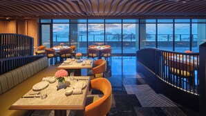 A restaurant with a view of a snowy landscape through large windows. Tables are set with plates, glasses, and flowers. The ceiling has a wavy design, and there is a pool in the foreground. - Conrad Altay Hemu (Burqin)