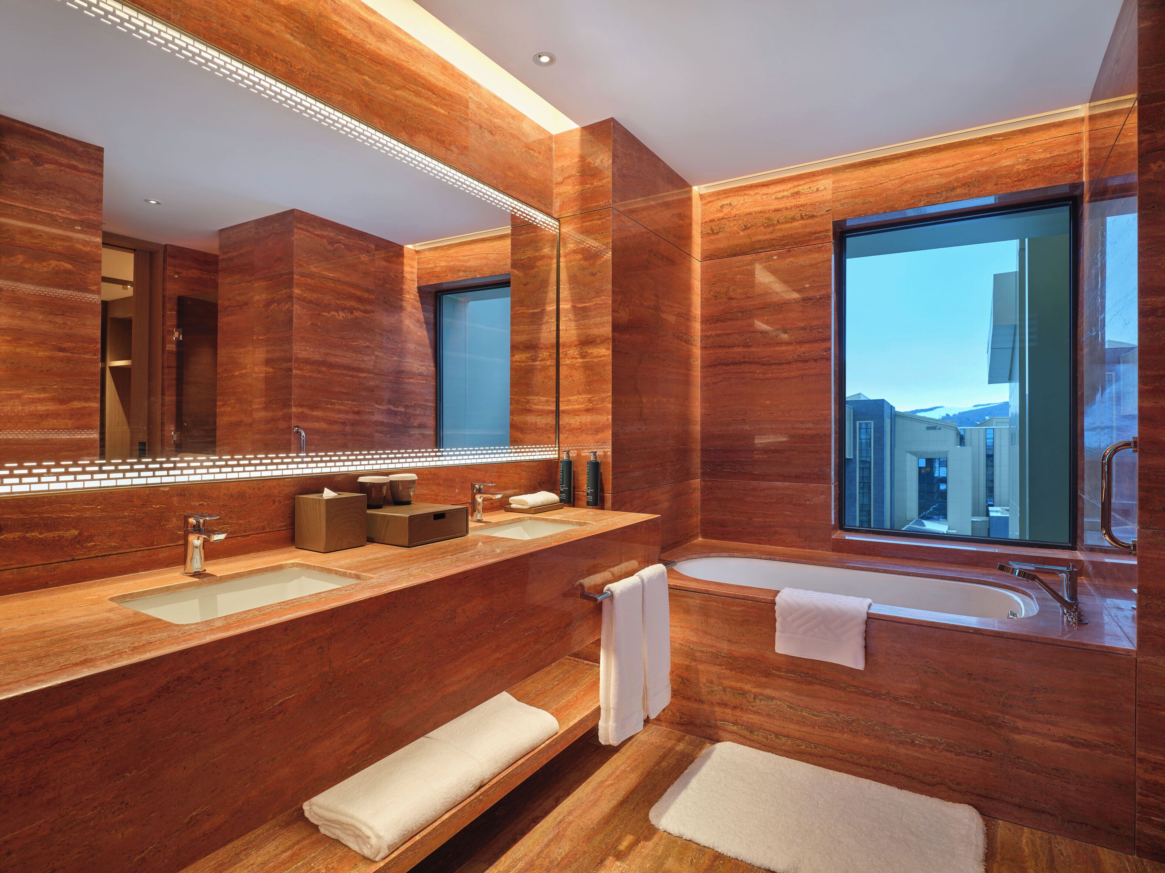 A modern bathroom with a large mirror, two sinks, a bathtub, and a window with a city view.