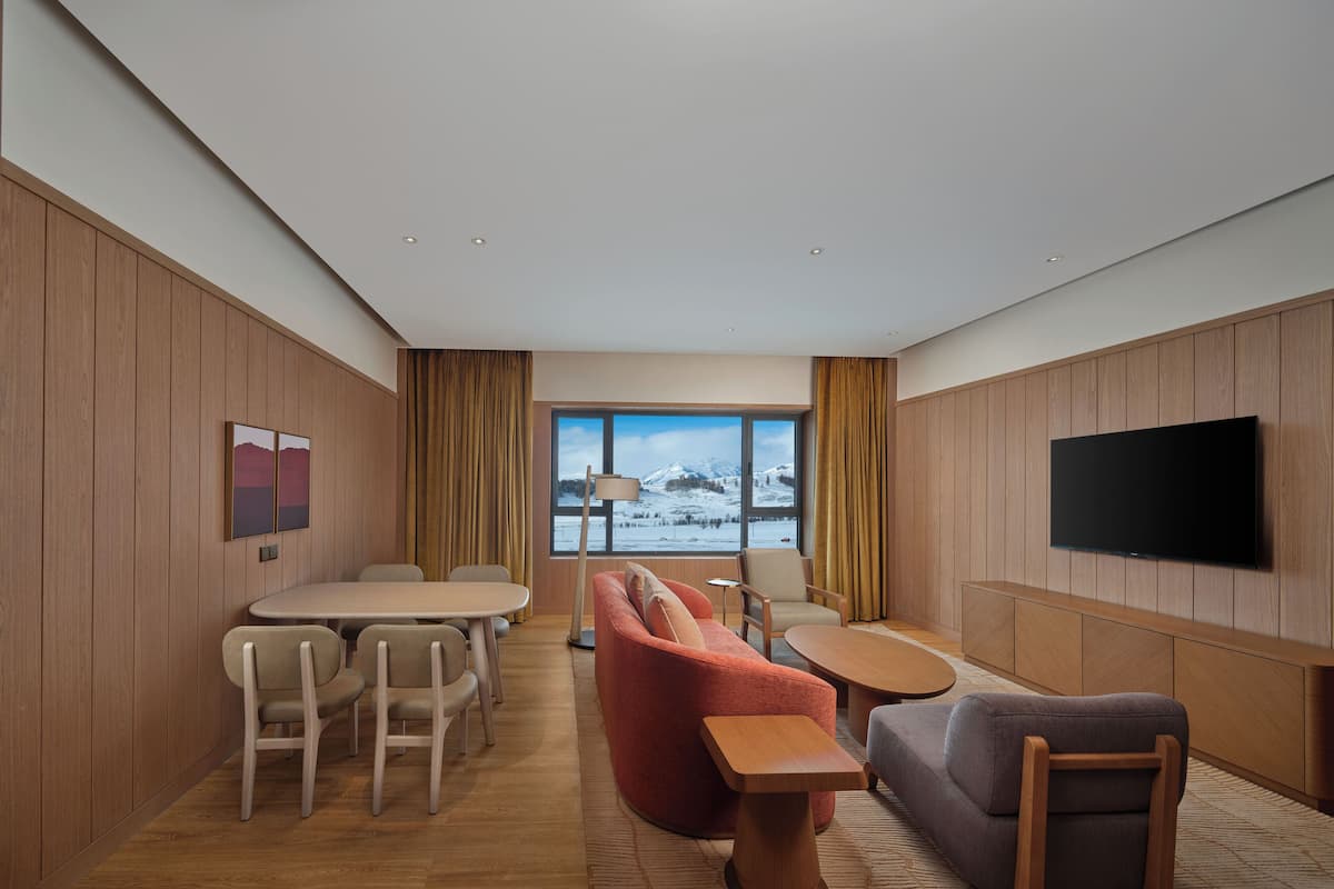 A modern hotel room with a wooden paneled wall, a flat-screen TV, a dining area with a table and chairs, a sofa, and a view of a snowy landscape through a large window.