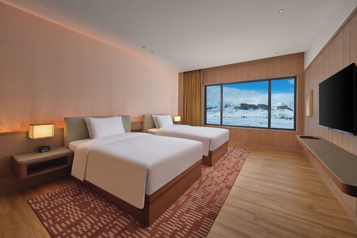 A hotel room with two beds, a large window with a snowy mountain view, a flat-screen TV, and wooden paneling.