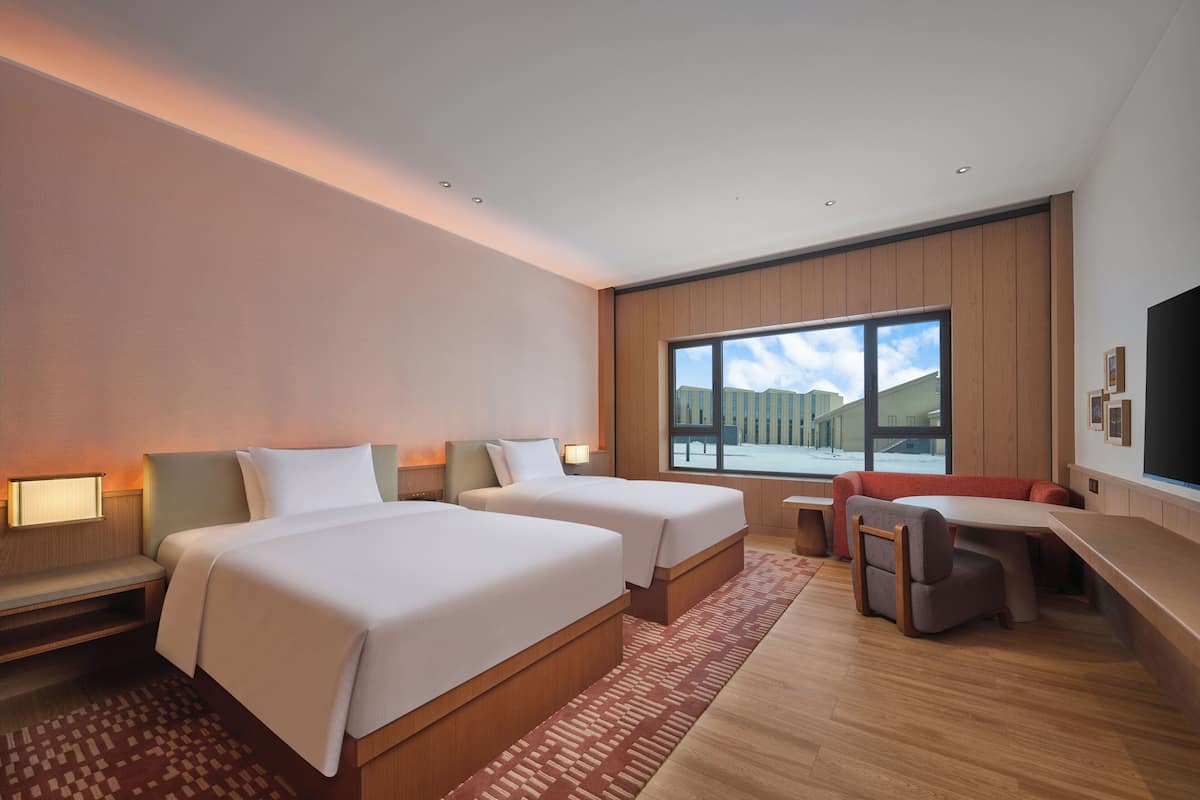 A hotel room with two beds, a desk, a chair, and a large window offering a view of buildings.