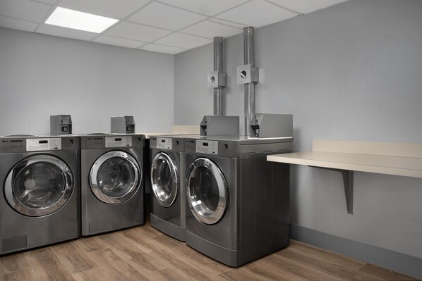A laundry room with three front-loading washing machines, a built-in shelf, and a grey wall. - Spark By Hilton Northborough (Northborough)