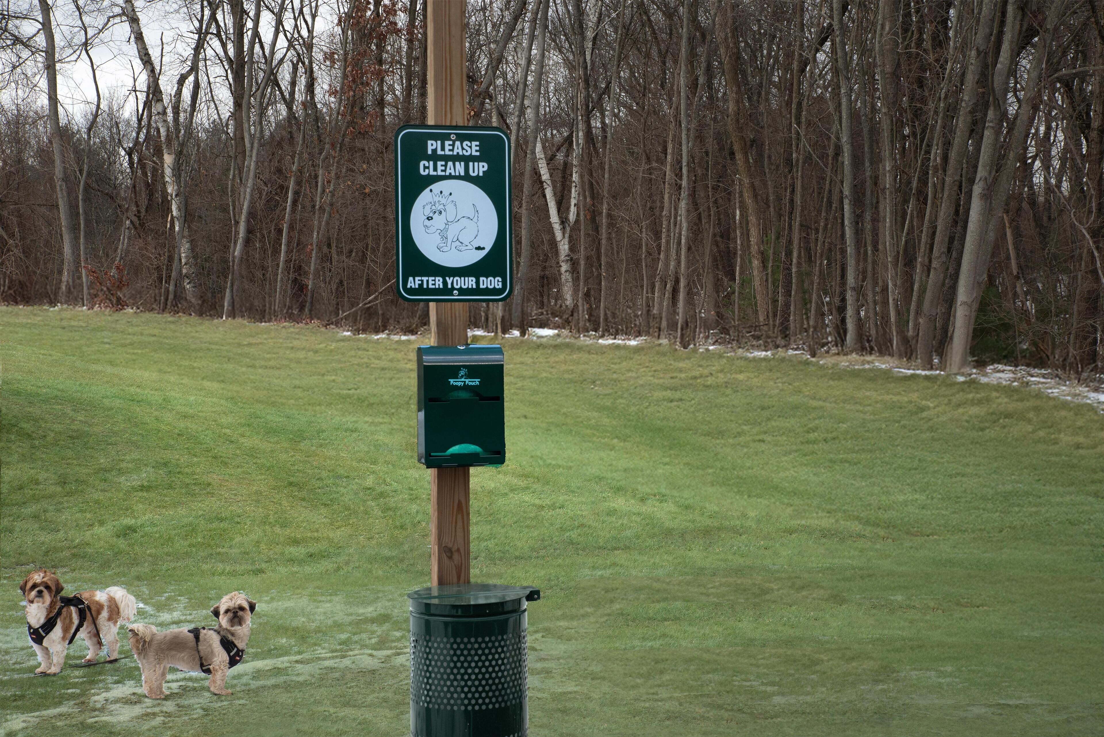 A sign instructing to clean up after dogs, a trash can, and two dogs on a grassy field.