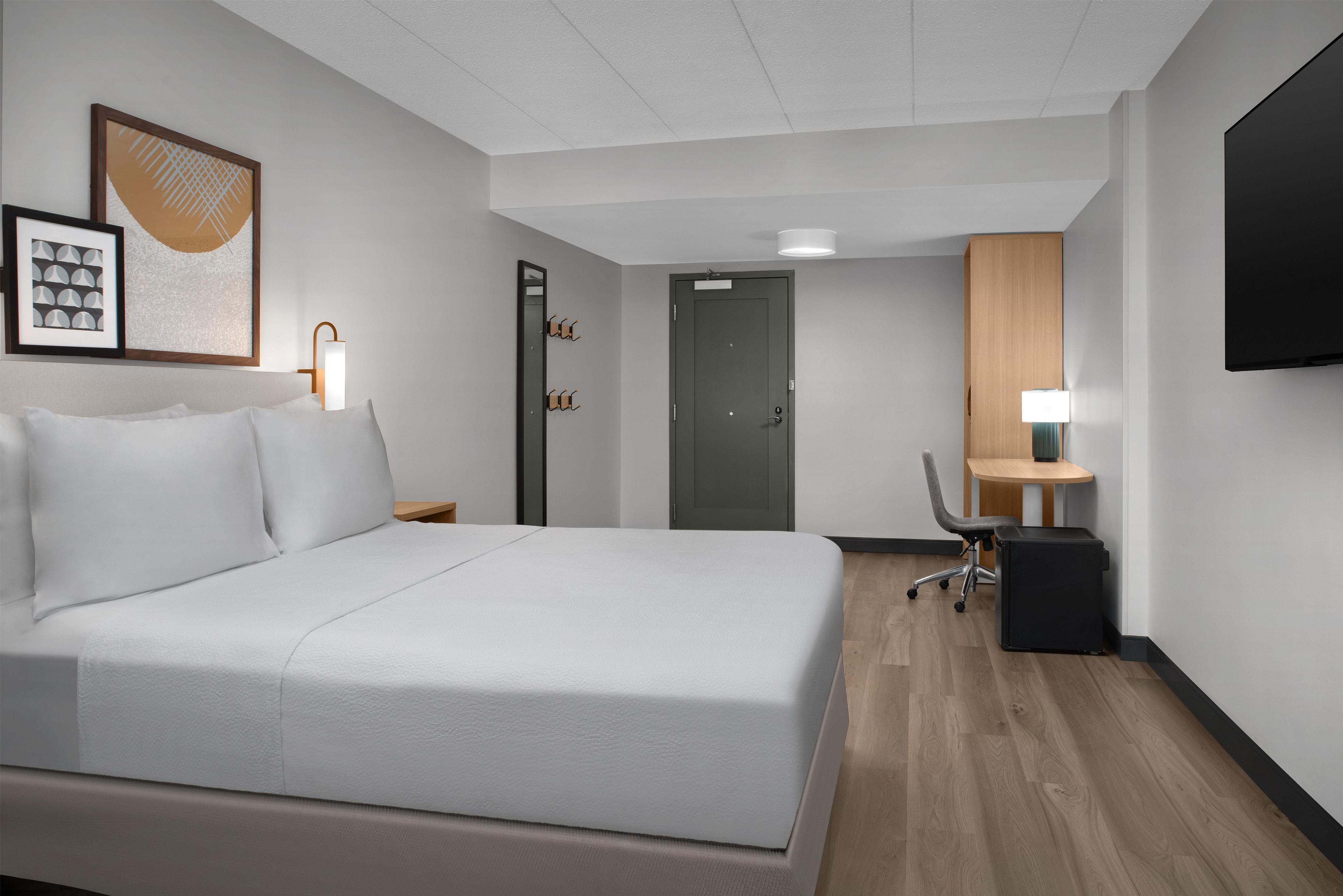 Ein Hotelzimmer mit einem großen Bett, einem Schreibtisch mit Stuhl, einem an der Wand befestigten Flachbildfernseher und einer Tür mit Spiegel.