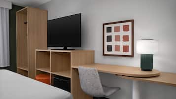 Habitación de hotel con cama, escritorio, silla, televisor, un cuadro enmarcado y lámpara.