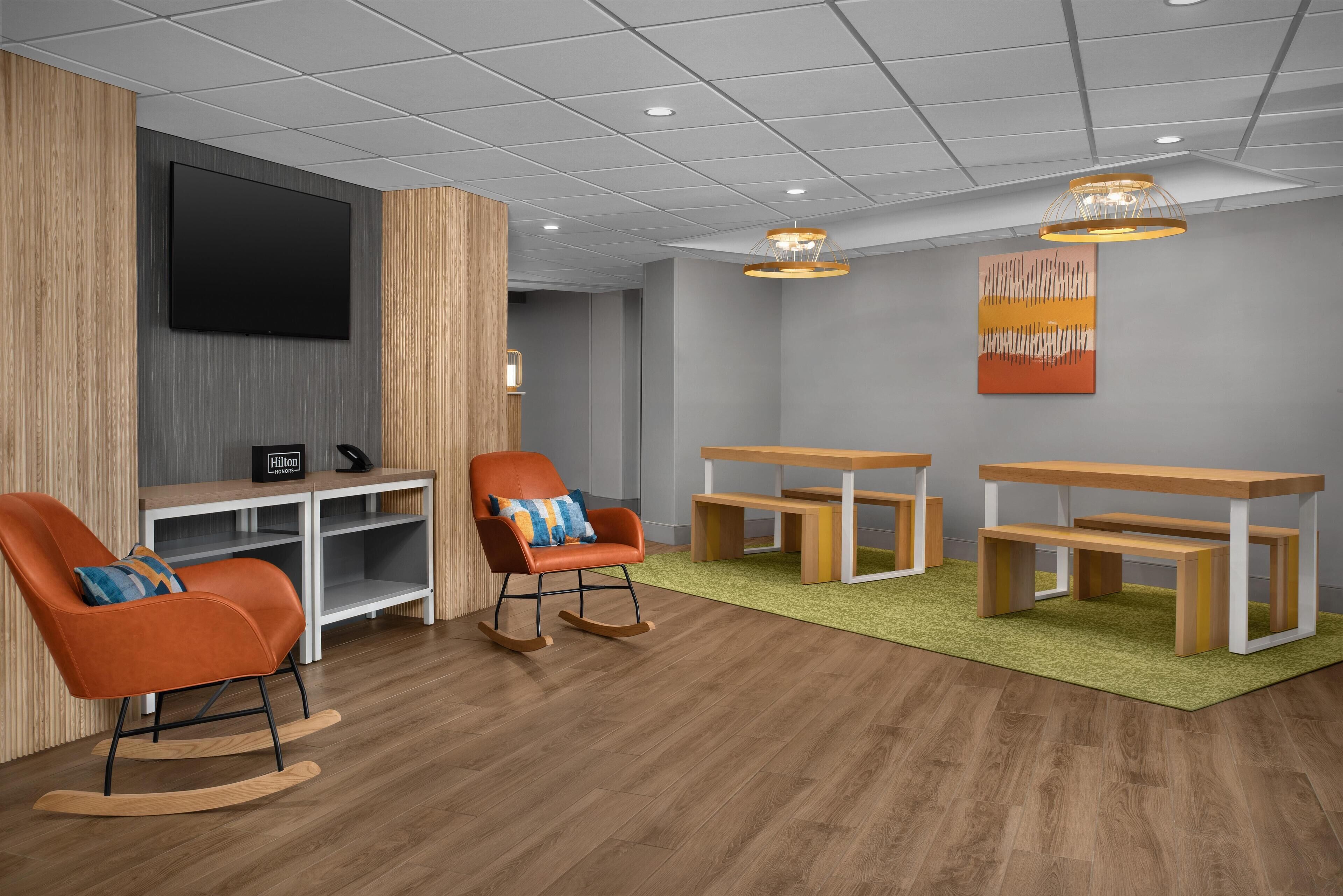 A modern hotel lobby with a flat-screen TV, rocking chairs, a wooden table, and a green carpet.