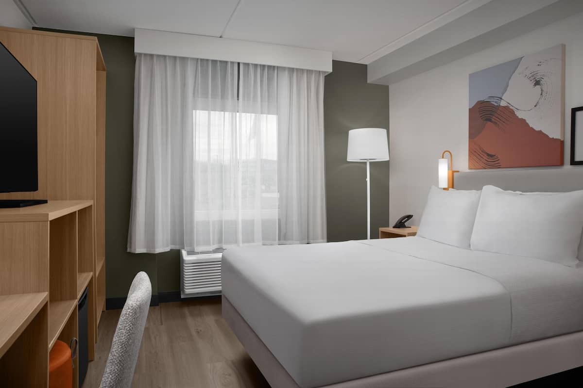 Een hotelkamer met een groot bed, een bureau met stoel, een staande lamp en een raam met dunne gordijnen.