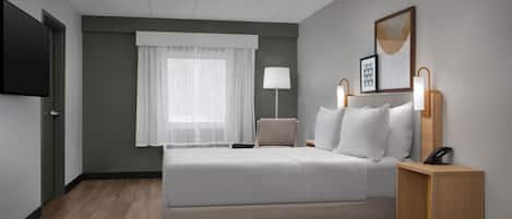 Una habitación de hotel moderna con una cama grande, un televisor de pantalla plana, una mesita de noche con lámpara y un cuadro enmarcado en la pared.