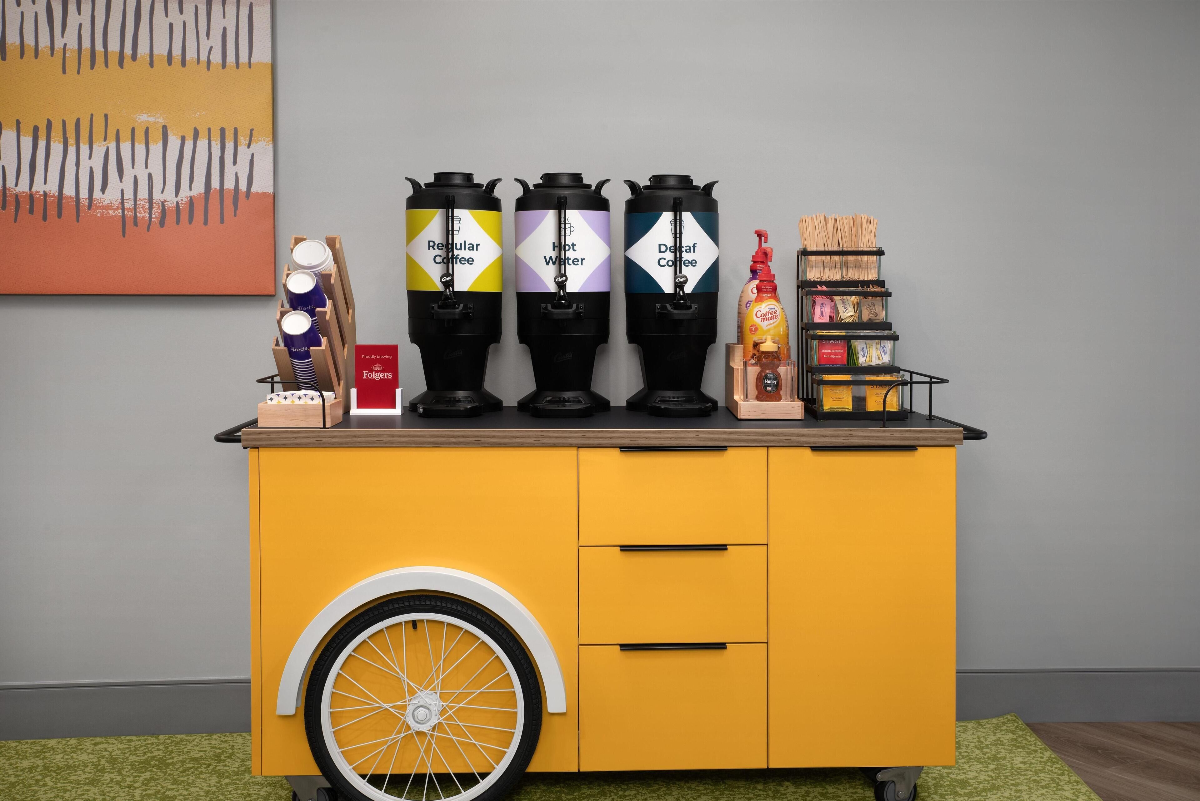 A yellow cart with three coffee dispensers labeled "Regular Coffee," "Hot Water," and "Decaf Coffee," and a Folgers coffee box.