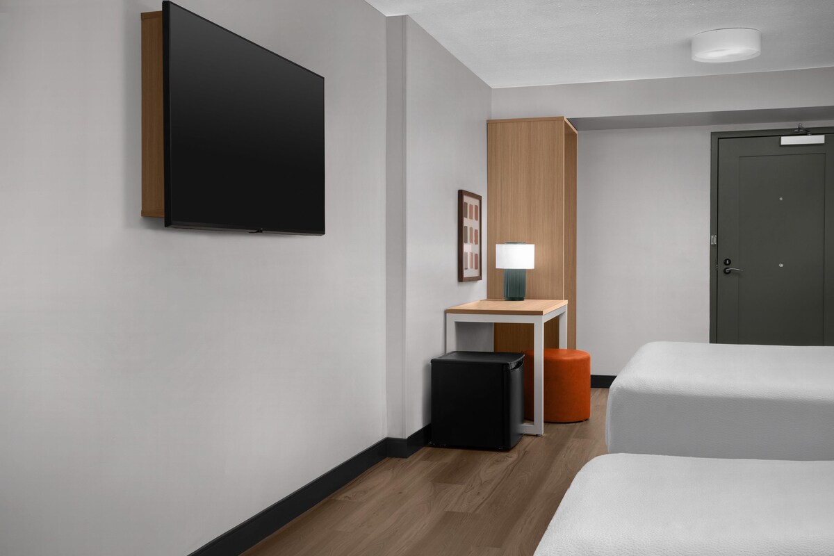 Camera d'albergo con televisione a schermo piatto montata a parete, un piccolo tavolo con lampada, un cestino e due letti.