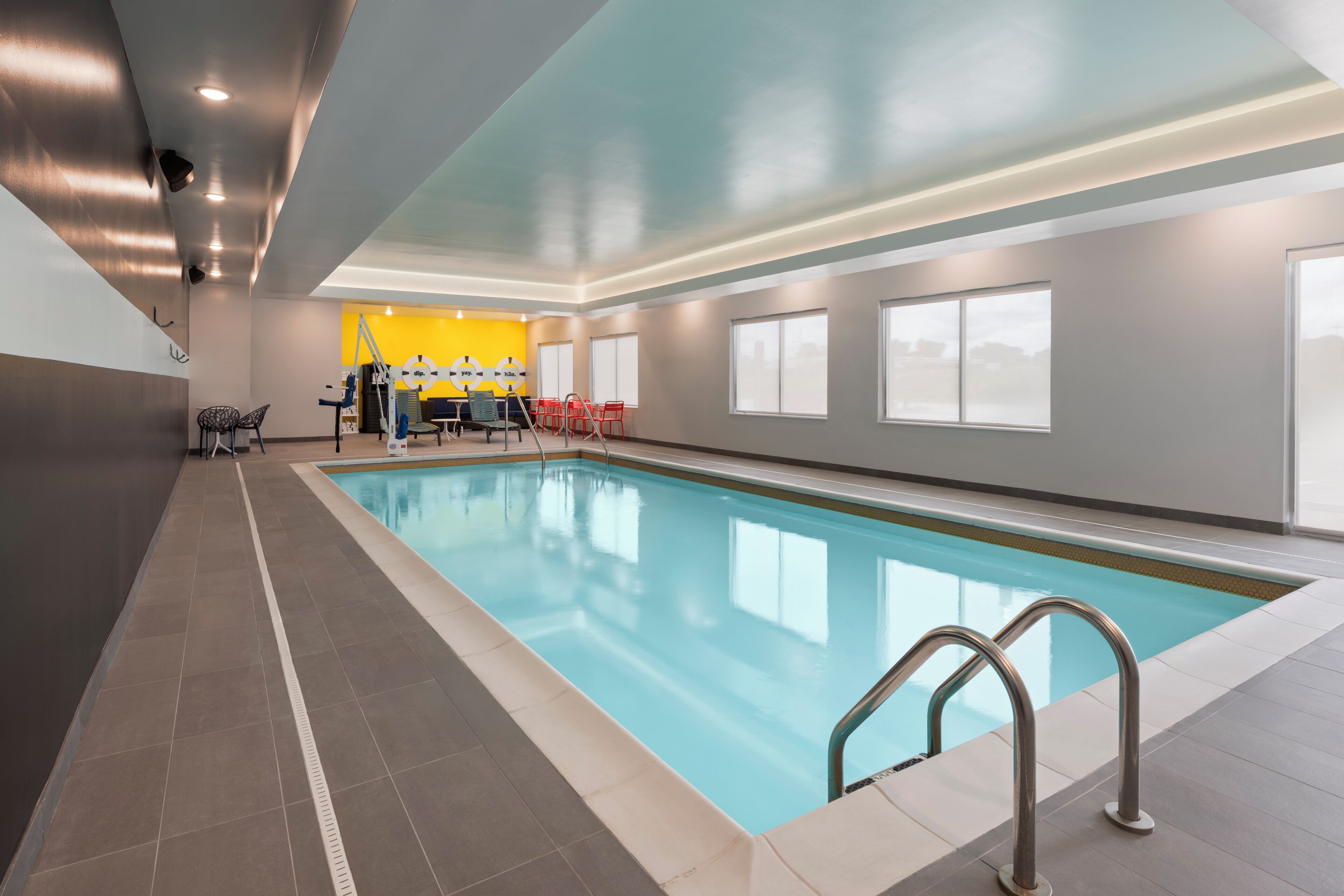 Une piscine avec une terrasse carrelée, un vestiaire équipé de bancs et un mur jaune orné d’un logo.