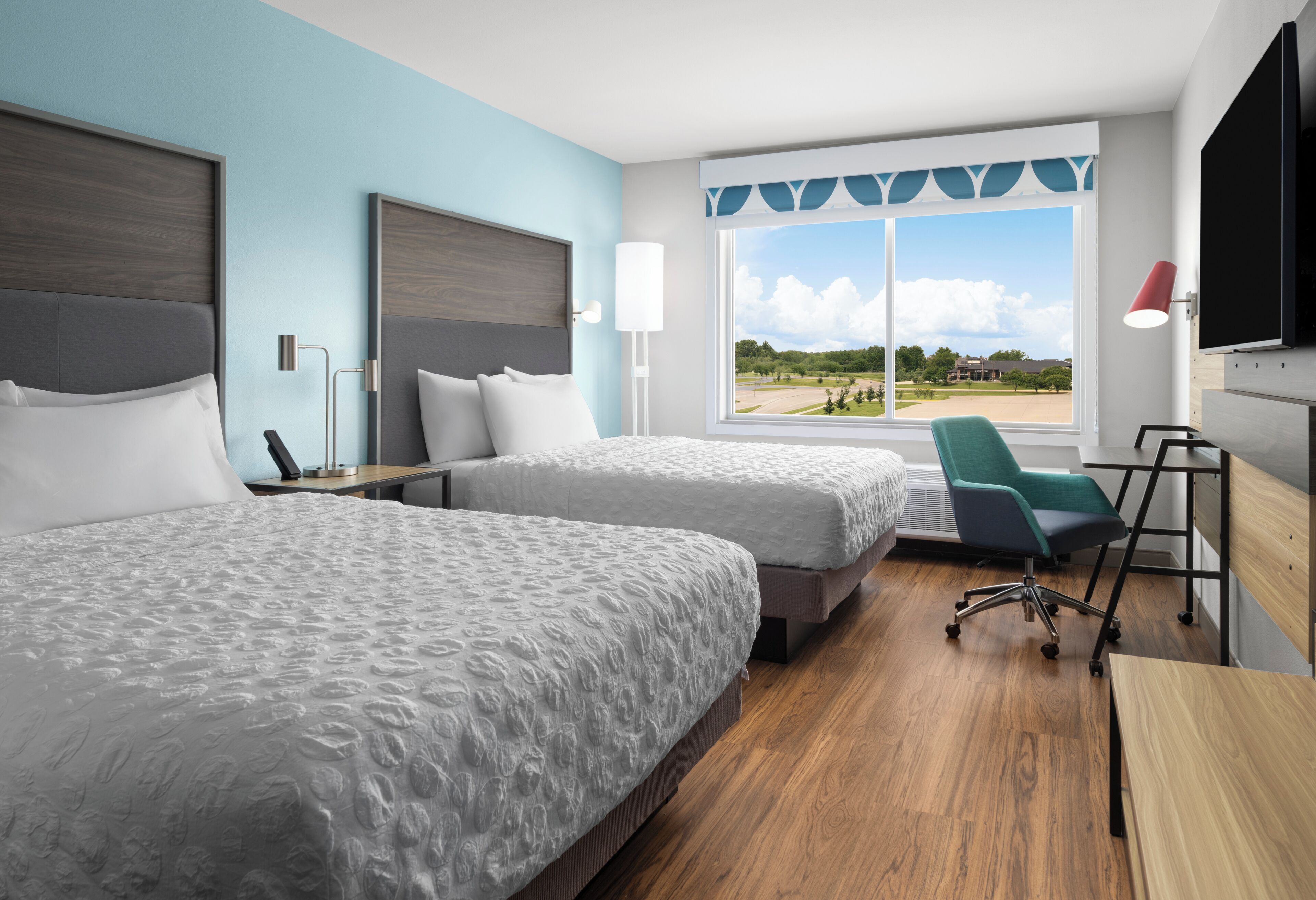 Une chambre d’hôtel avec deux lits, un bureau, une chaise, une télévision et une fenêtre donnant sur un immeuble et des arbres.