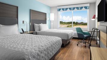 Une chambre d’hôtel avec deux lits, un bureau, une chaise, une télévision et une fenêtre donnant sur un immeuble et des arbres.