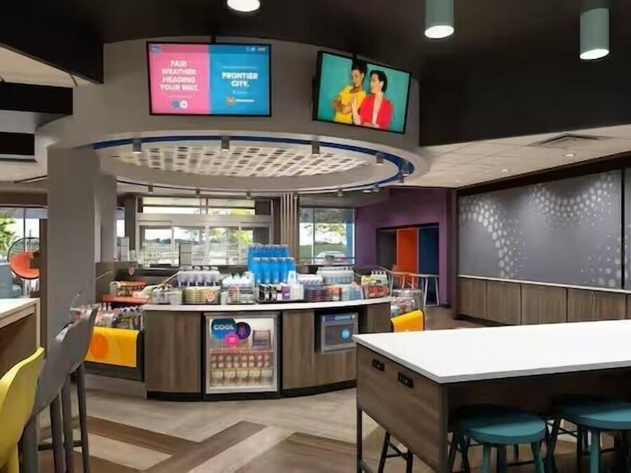 A modern cafe with a central counter, seating area, and two digital screens displaying advertisements.
