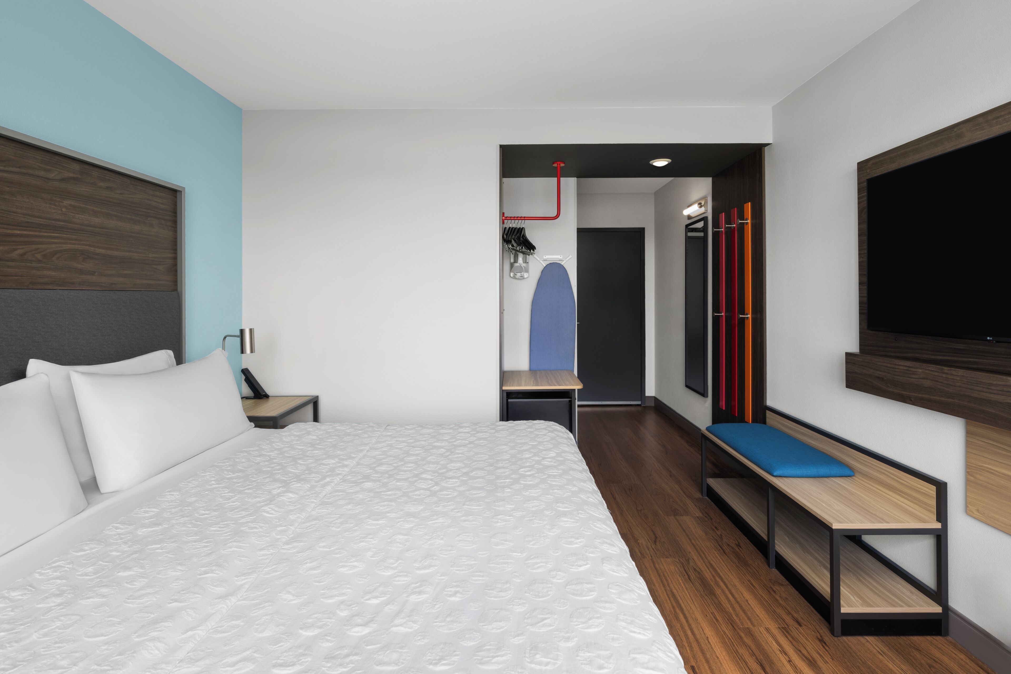 A modern hotel room with a large bed, a desk, a bench, and a TV mounted on the wall.