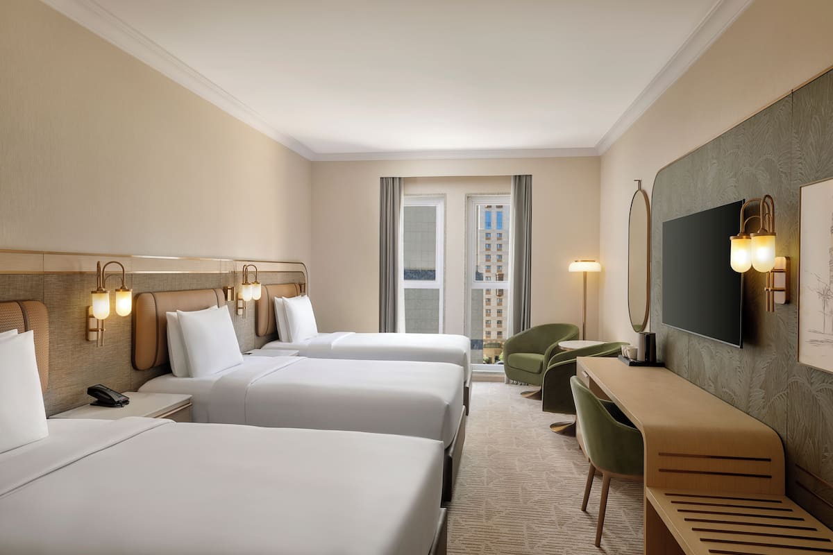 Habitación de hotel con tres camas, un escritorio, una silla, un televisor y un teléfono en la mesita de noche.