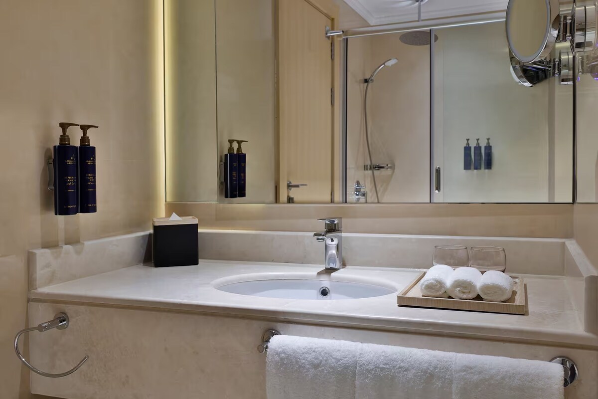 A bathroom with a large mirror, a sink, and a shower. There are toiletries on the wall and towels on the counter.