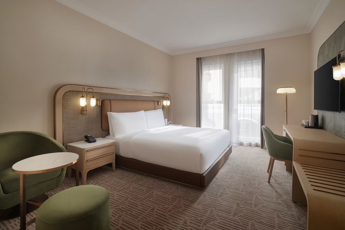 A hotel room with a large bed, a desk, two chairs, a nightstand, and a TV.