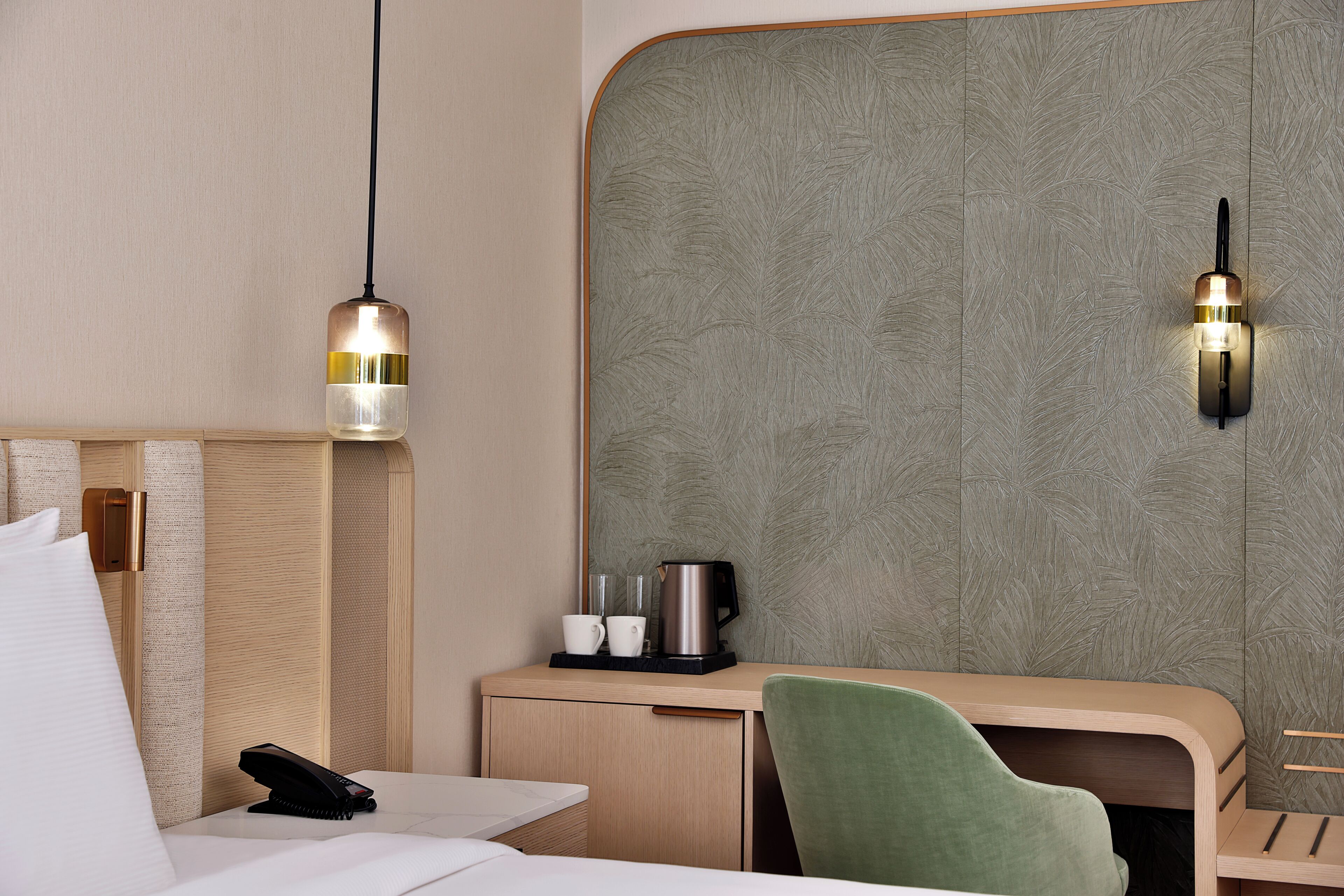 A hotel room with a bed, bedside table, green chair, and a desk with a kettle and cups.