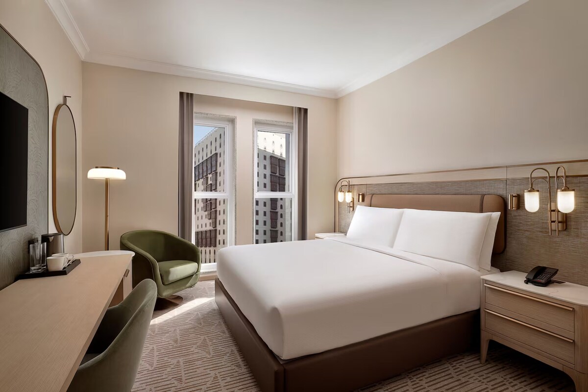 A hotel room with a large bed, a desk with a chair, a bedside table with a phone, and a large window with a city view.