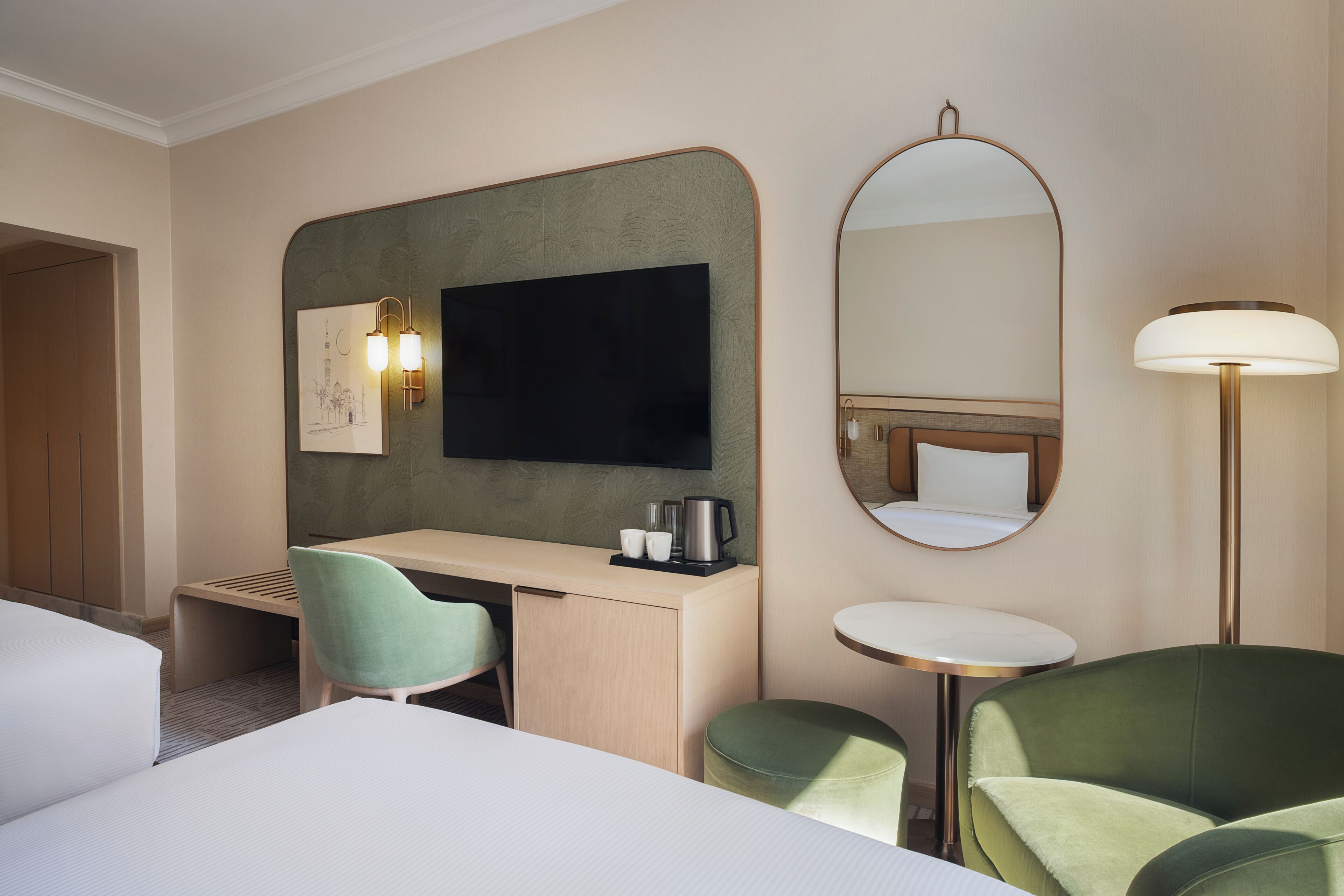 A hotel room with a bed, a desk, a chair, a round mirror, a bedside table, and a lamp.