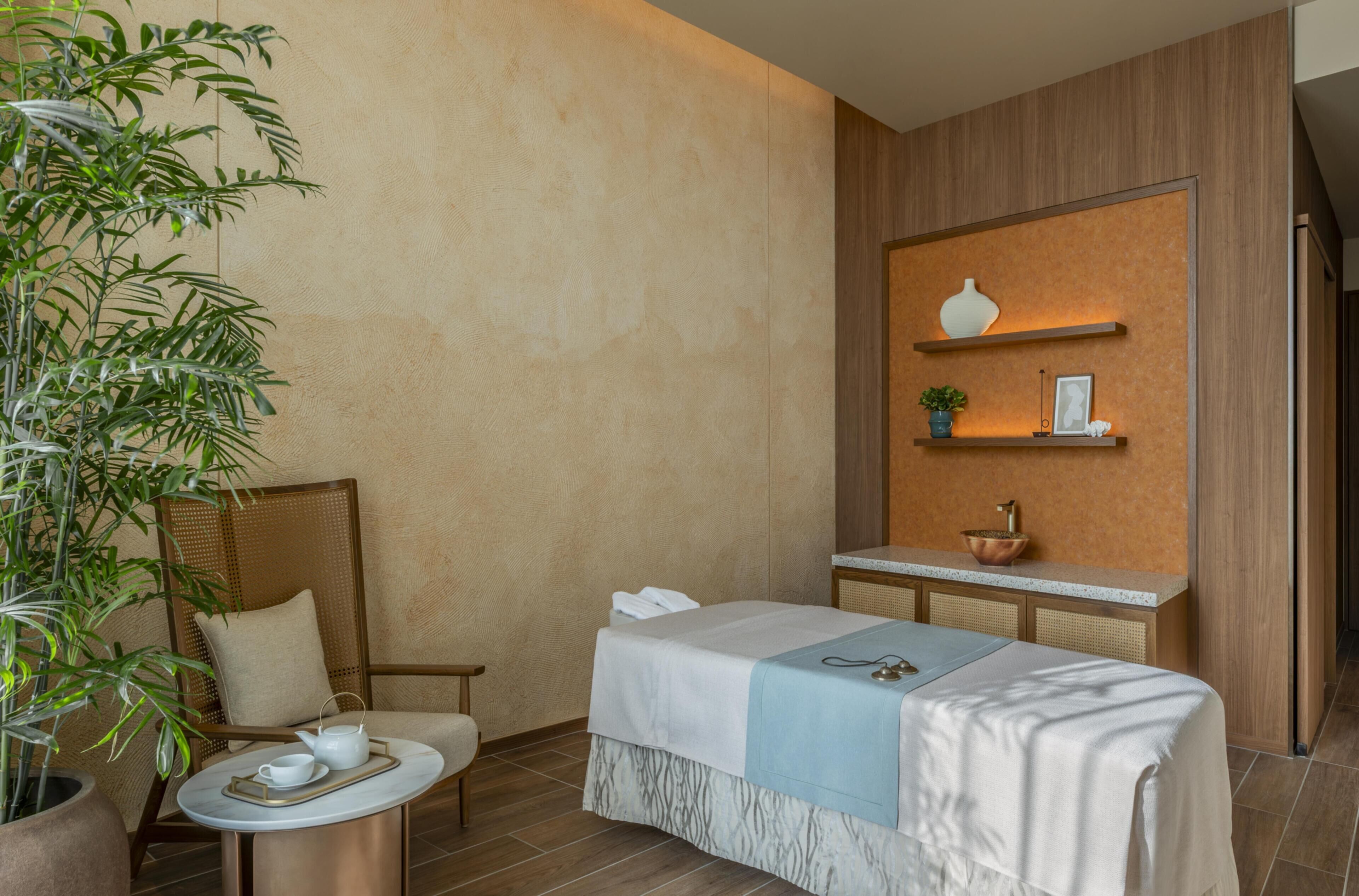 Un cuarto de spa con una camilla de masaje, una silla, una mesita con tetera y tazas, una planta en maceta y un estante de pared con objetos decorativos.