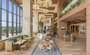 Eine großzügige Lobby mit hoher Decke, großen Fenstern und einer zentralen Sitzecke mit Tischen und Stühlen.