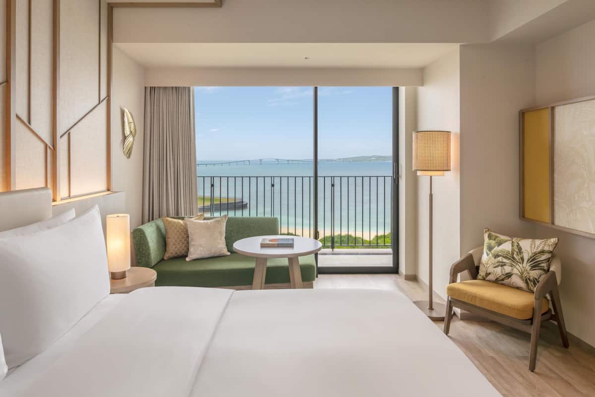 A hotel room with a large bed, a sofa, a small round table, a chair, a lamp, and a view of the sea and a bridge.
