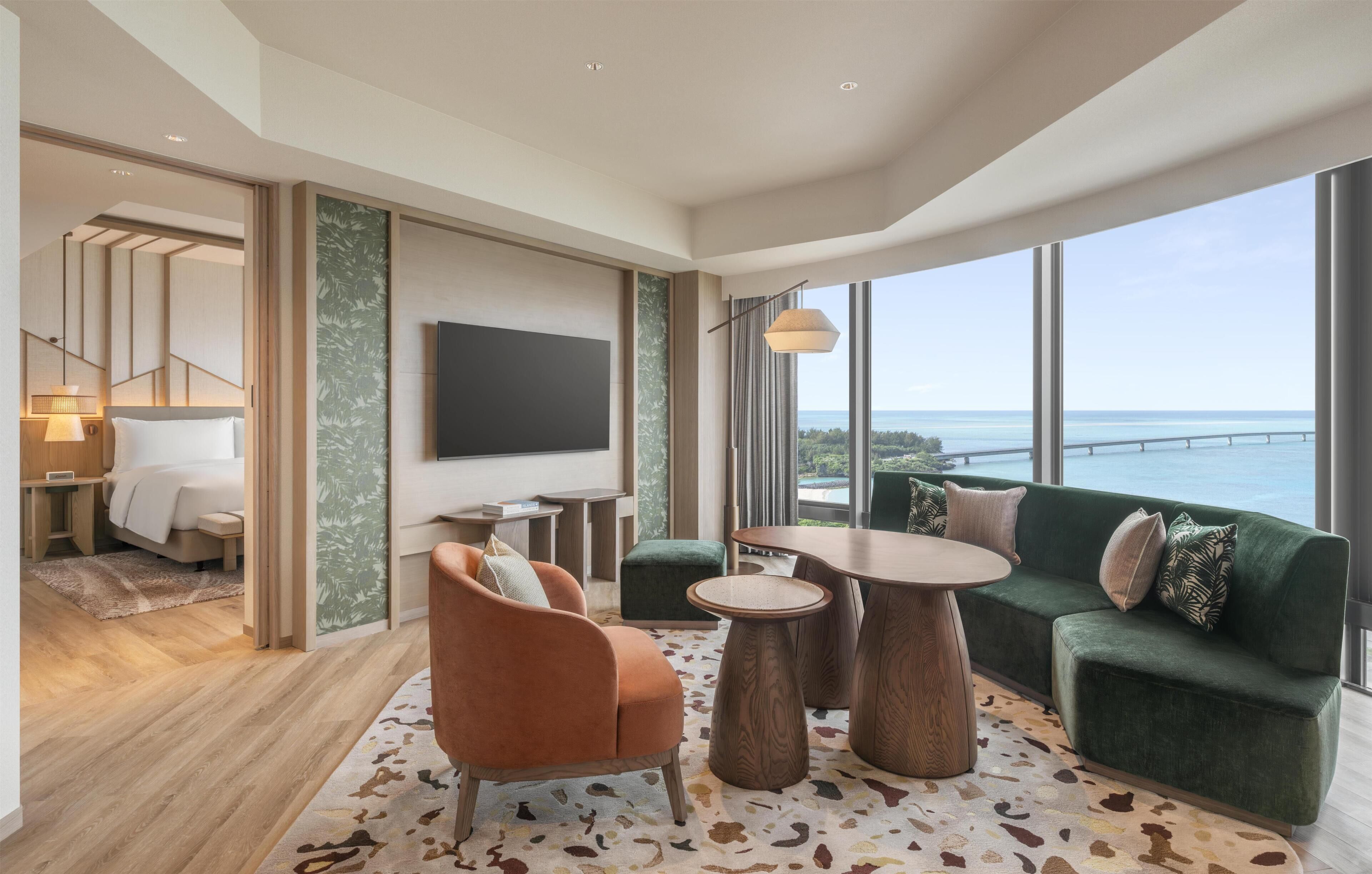 A modern hotel room with a large window, a green sofa, a wooden coffee table, and a flat-screen TV.
