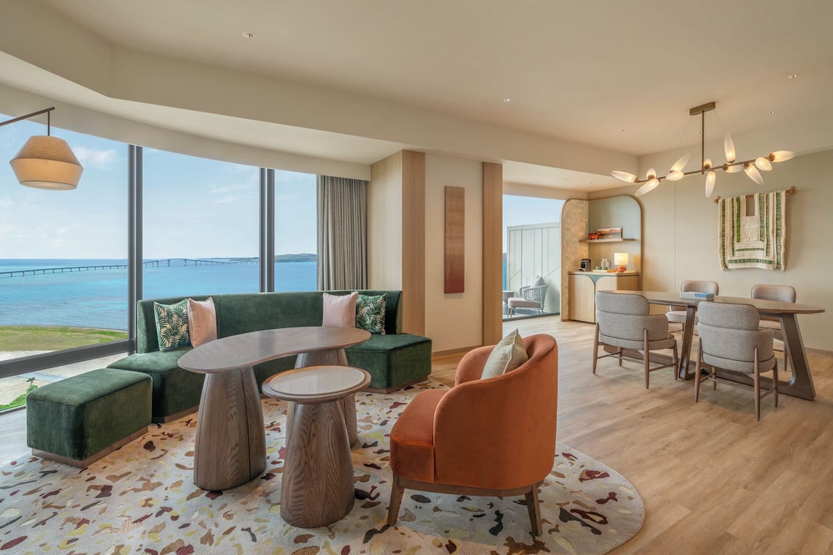 A modern hotel room with a large window, a green sofa, an orange armchair, a round table, and a dining area with chairs.