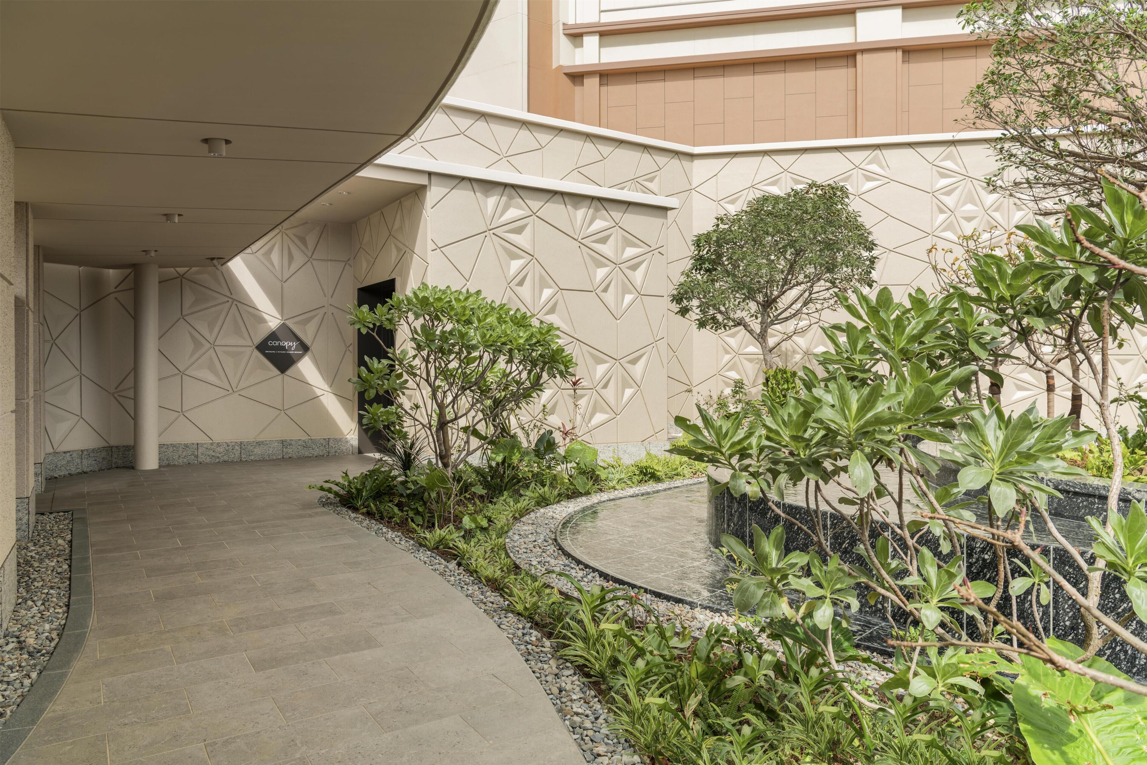 A curved pathway leading to a building entrance with geometric wall patterns and a variety of greenery.