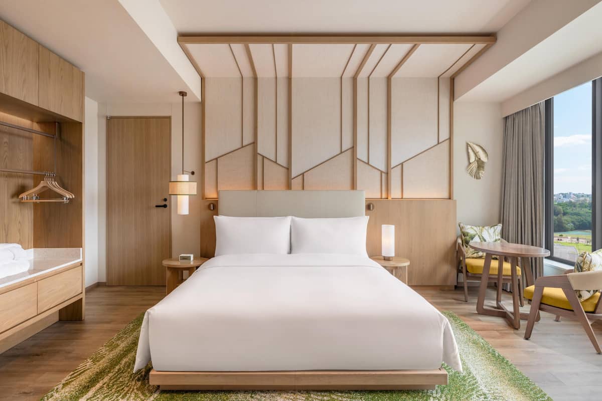 A modern hotel room with a large bed, wooden wardrobe, bedside tables, a small round table, and a view of the city.