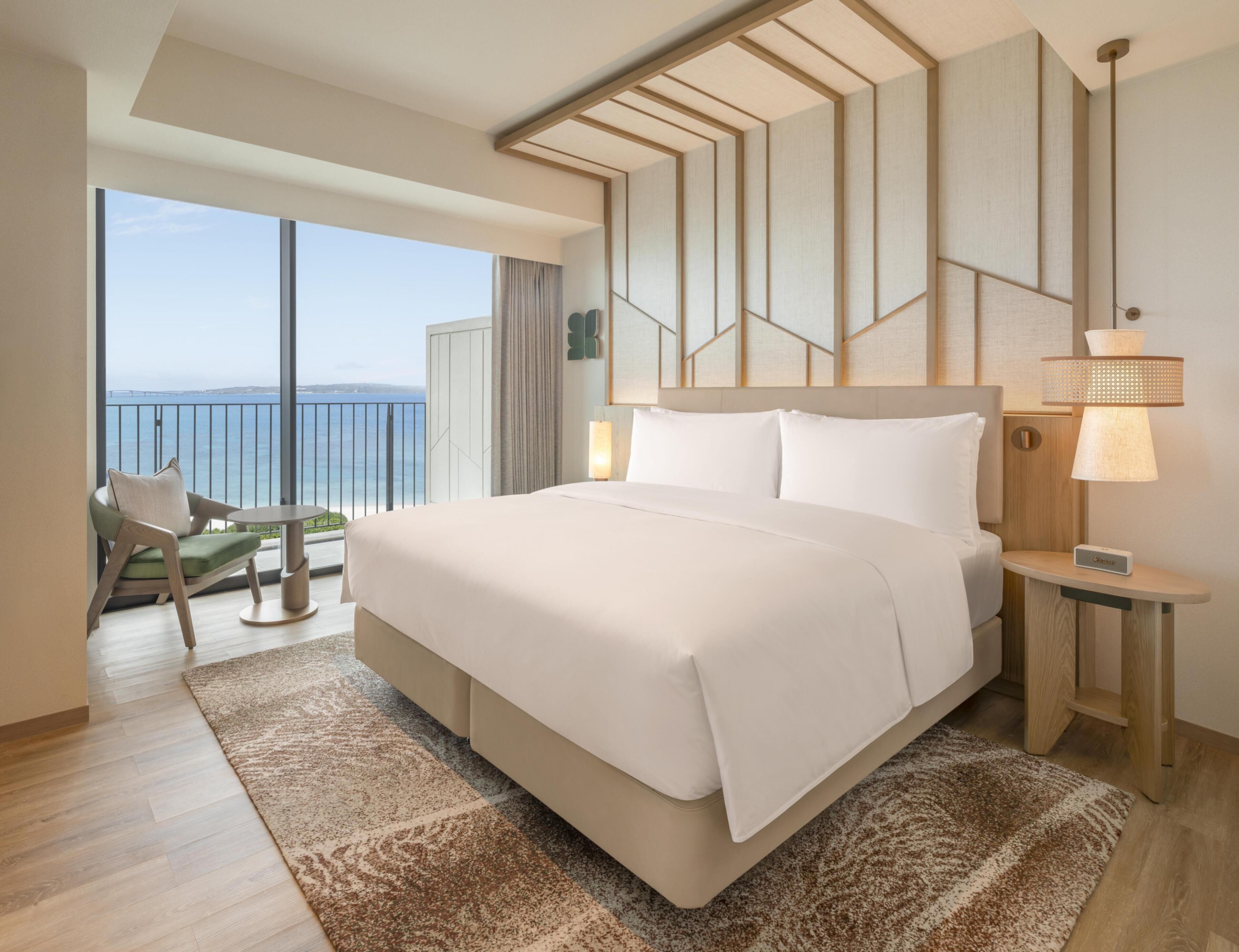 A modern hotel room with a large bed, a view of the sea, a balcony with a chair, and a nightstand with a lamp.
