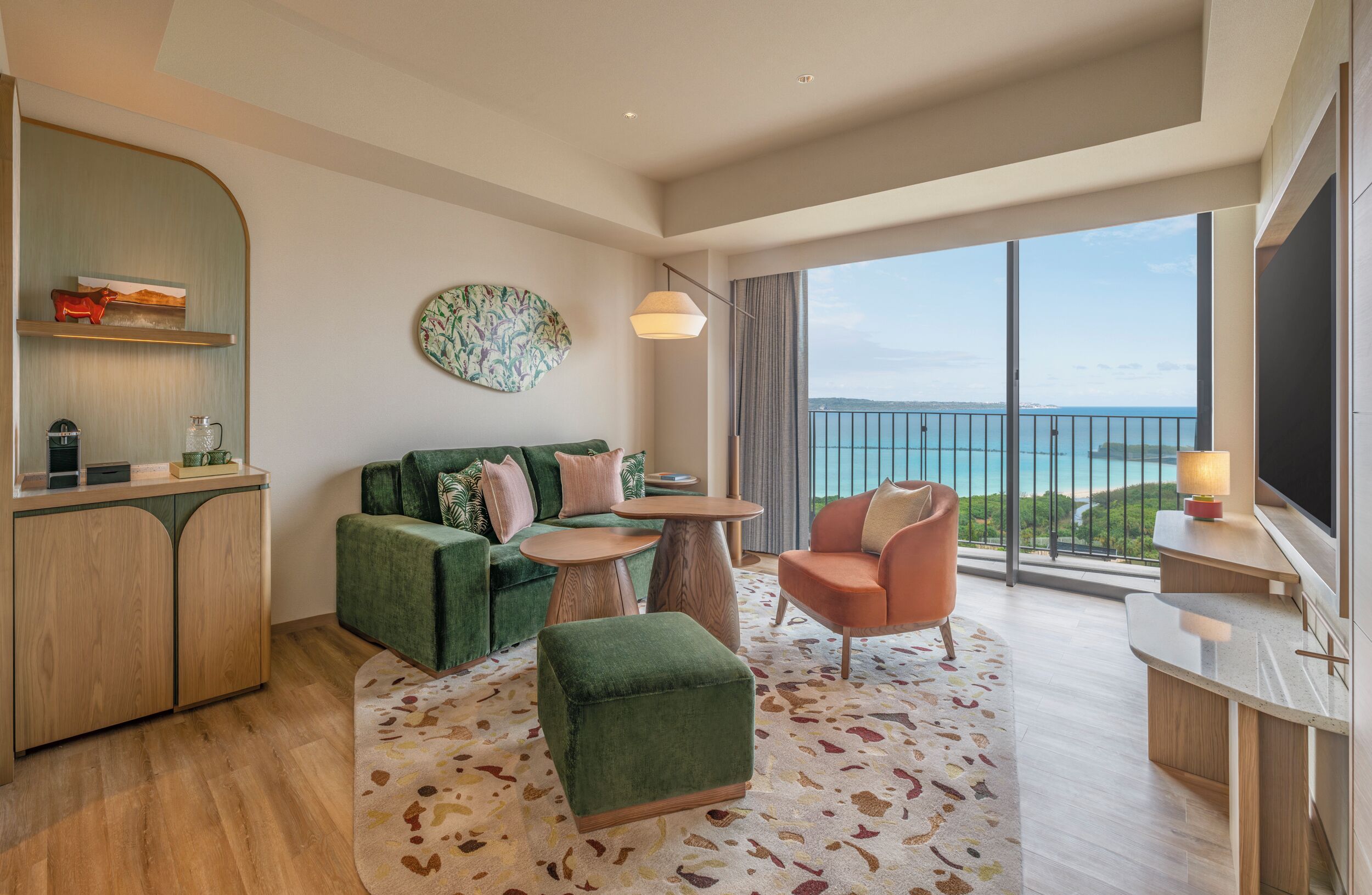 A modern hotel room with a green sofa, a small round table, a wooden cabinet, and a large window with a view of the sea.
