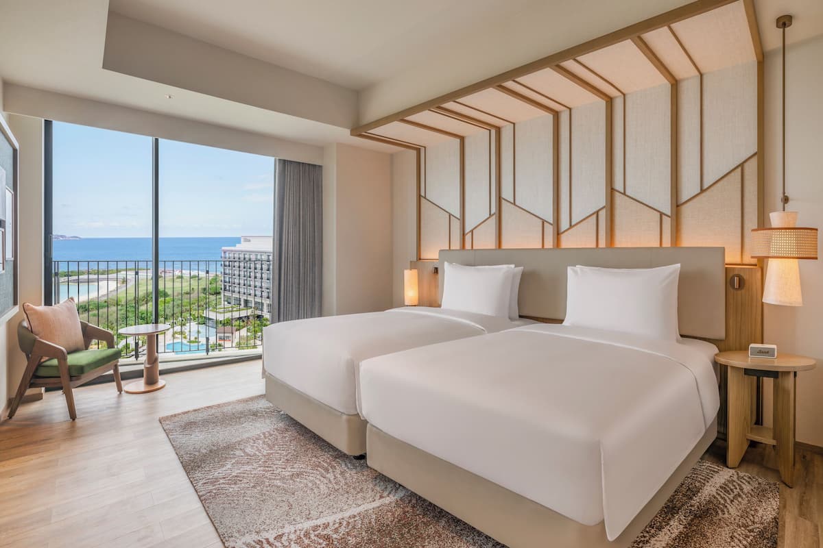 A hotel room with two beds, a view of the ocean, a chair, a small table, and a lamp.