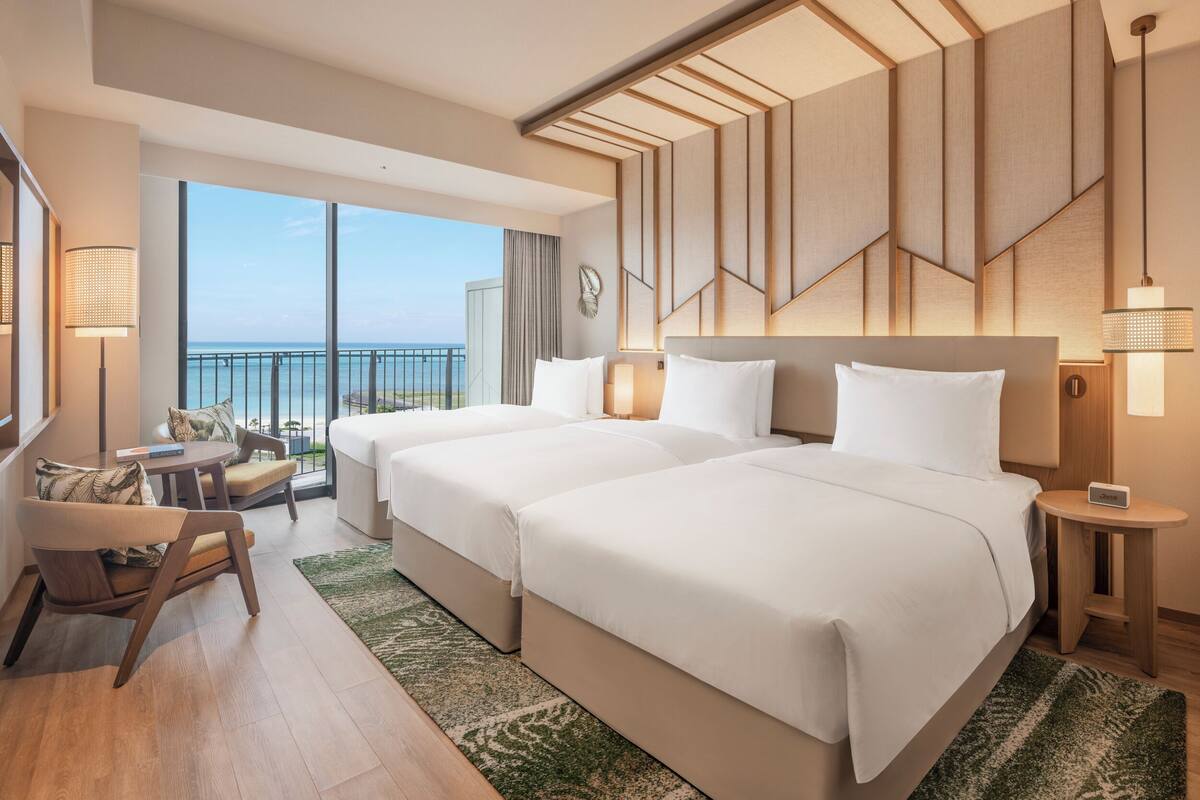 A hotel room with two beds, a balcony with a view, and a seating area with a table and chairs.