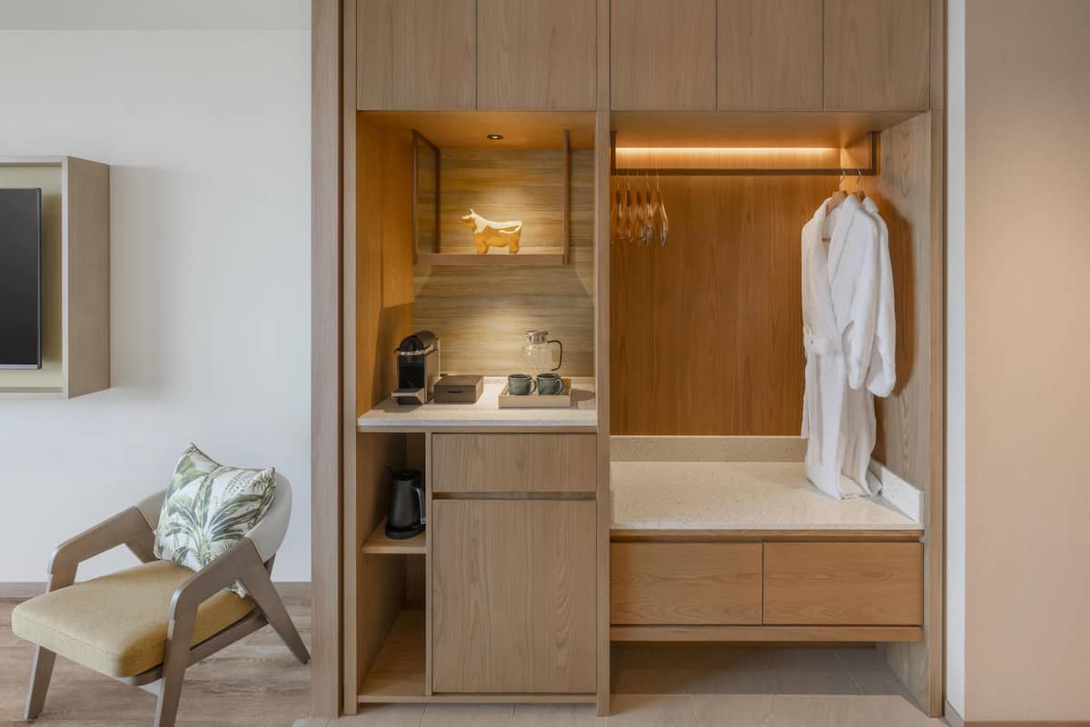 A compact room with a built-in desk, a seating area, and a walk-in closet with a mirror and hanging clothes.