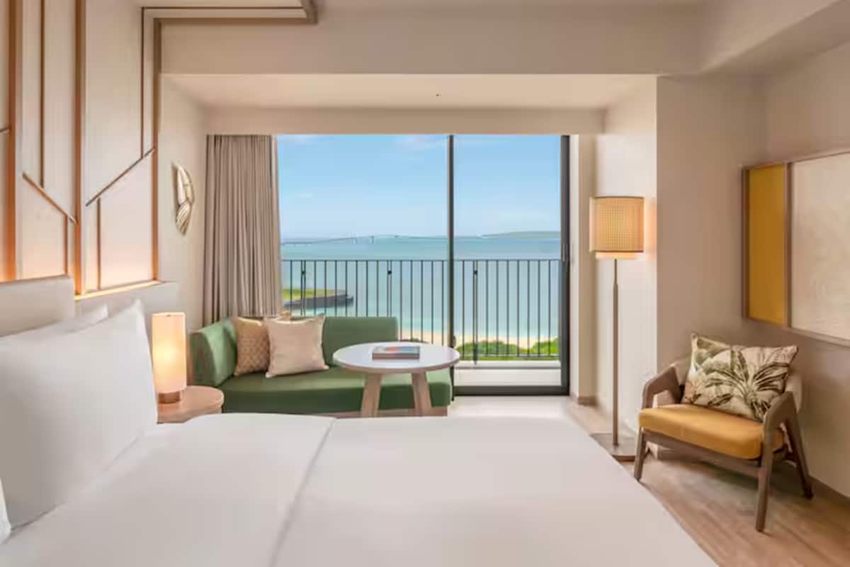 A hotel room with a large bed, a green sofa, a small round table, a chair, and a balcony with a view of the sea.